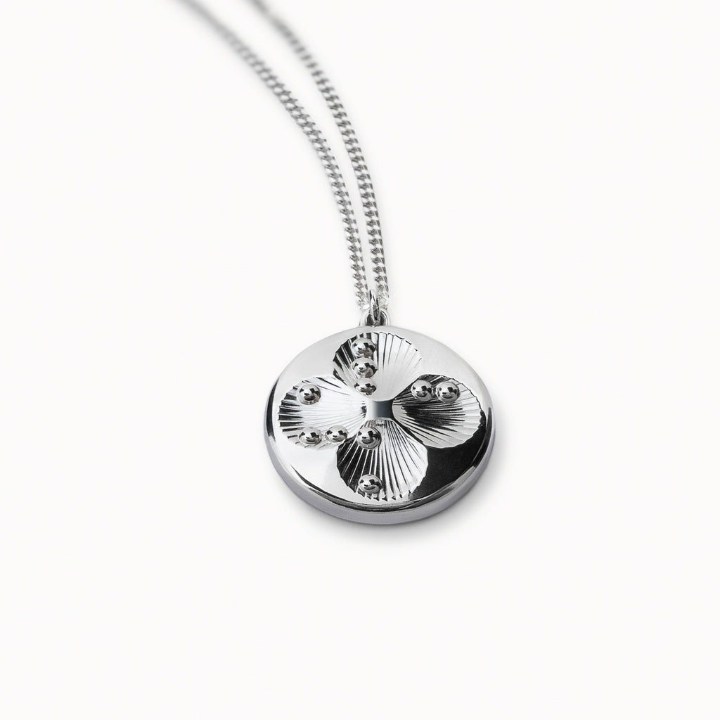 Lucky Dots smooth silver pendant with four shallow circular dips forming a four-leaf clover, each marked with the Braille letters L, U, C, and K. Fine shell-like lines are engraved inside the dips, and the composition of the four roundels creates a subtle star-shaped pattern in the centre.